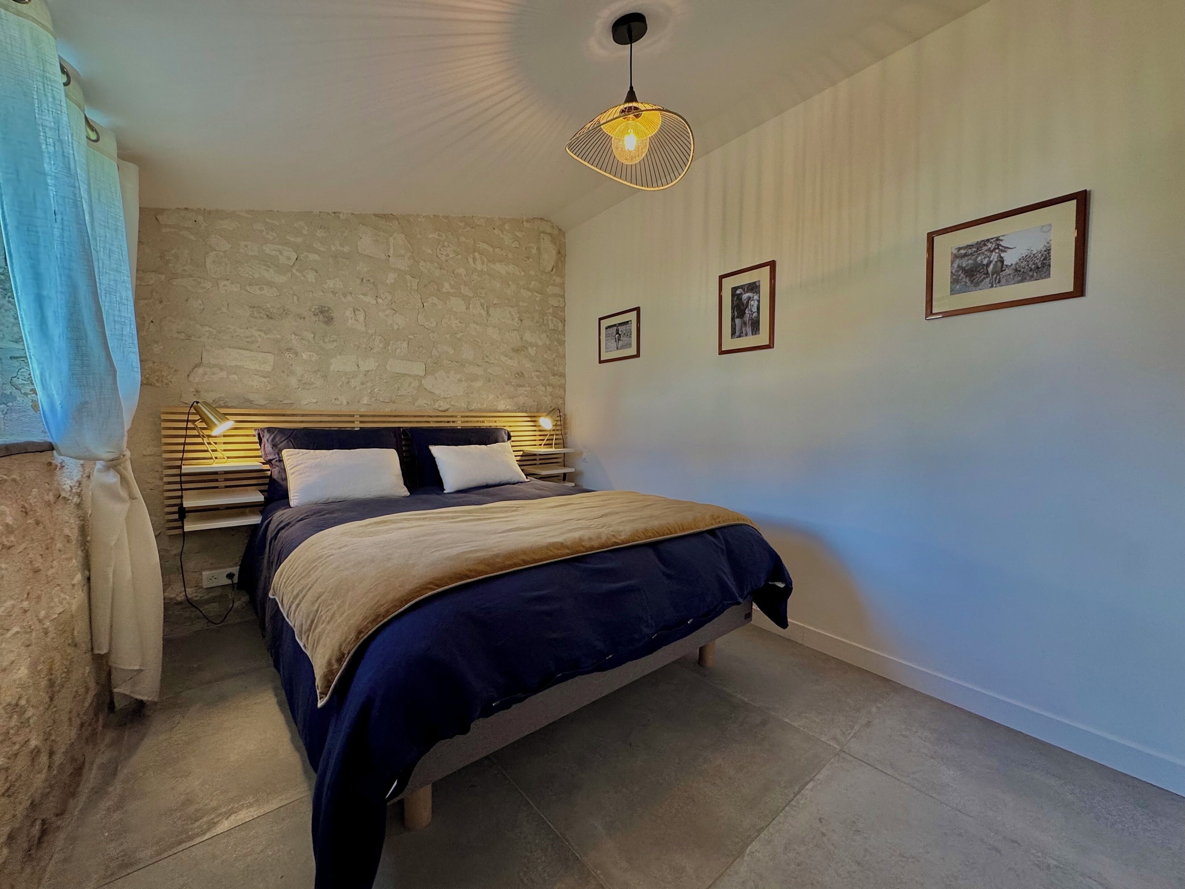 A bedroom with a stone wall, a bed with a headboard, a bedside table with a lamp, and framed pictures on the wall.