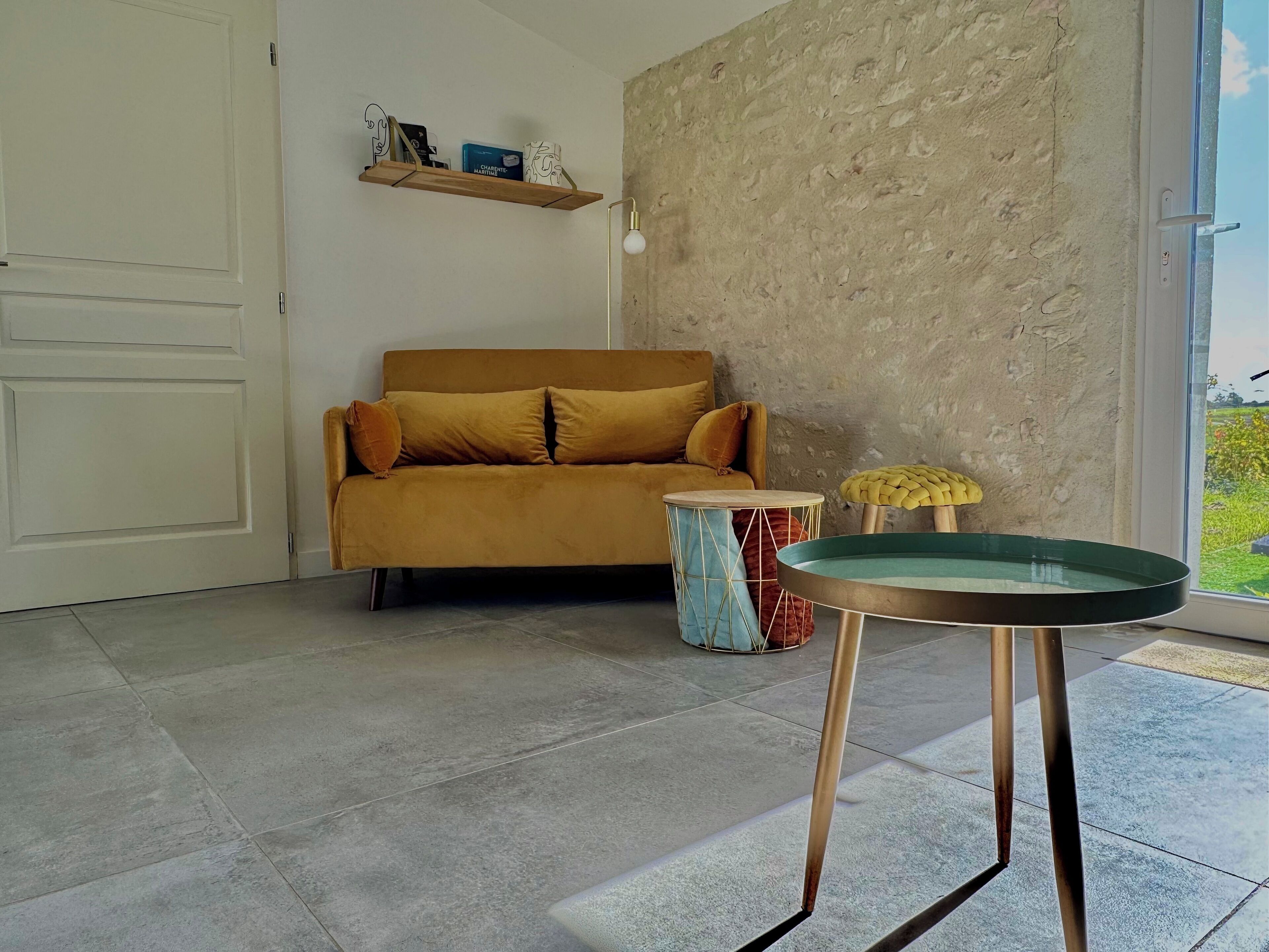 A living room with a sofa, a round table, and a drum. There is a shelf with items on the wall and a door in the background.