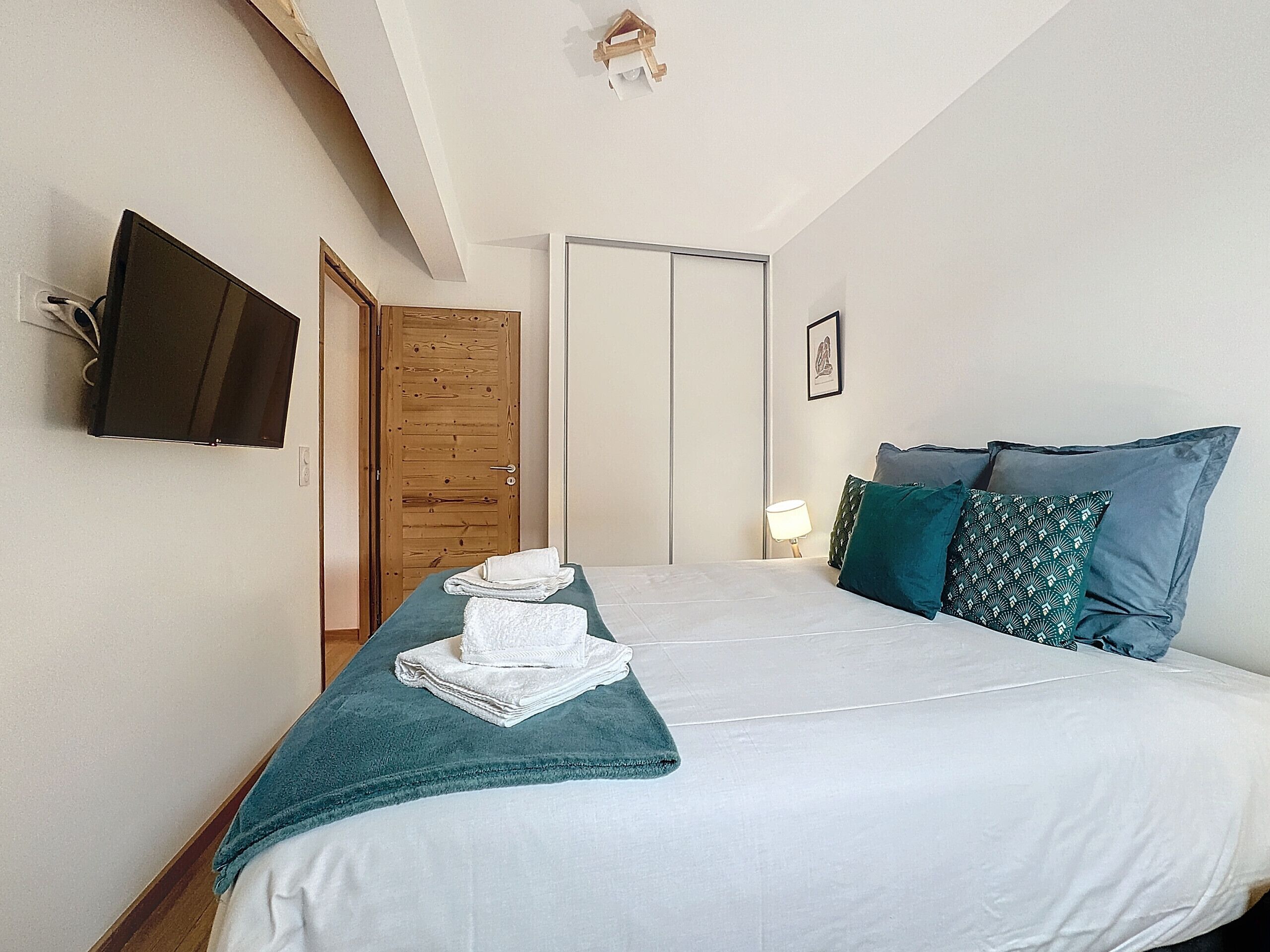 A hotel room with a bed, a TV mounted on the wall, a wooden door, and a bedside table with a lamp.