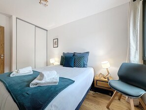A neatly made bed with a blue and white duvet, two pillows, and a folded towel. There is a bedside table with a lamp and a chair. - Les 3 Mousquetons- appartement- 6,8 Pers - Proche (Les Gets)