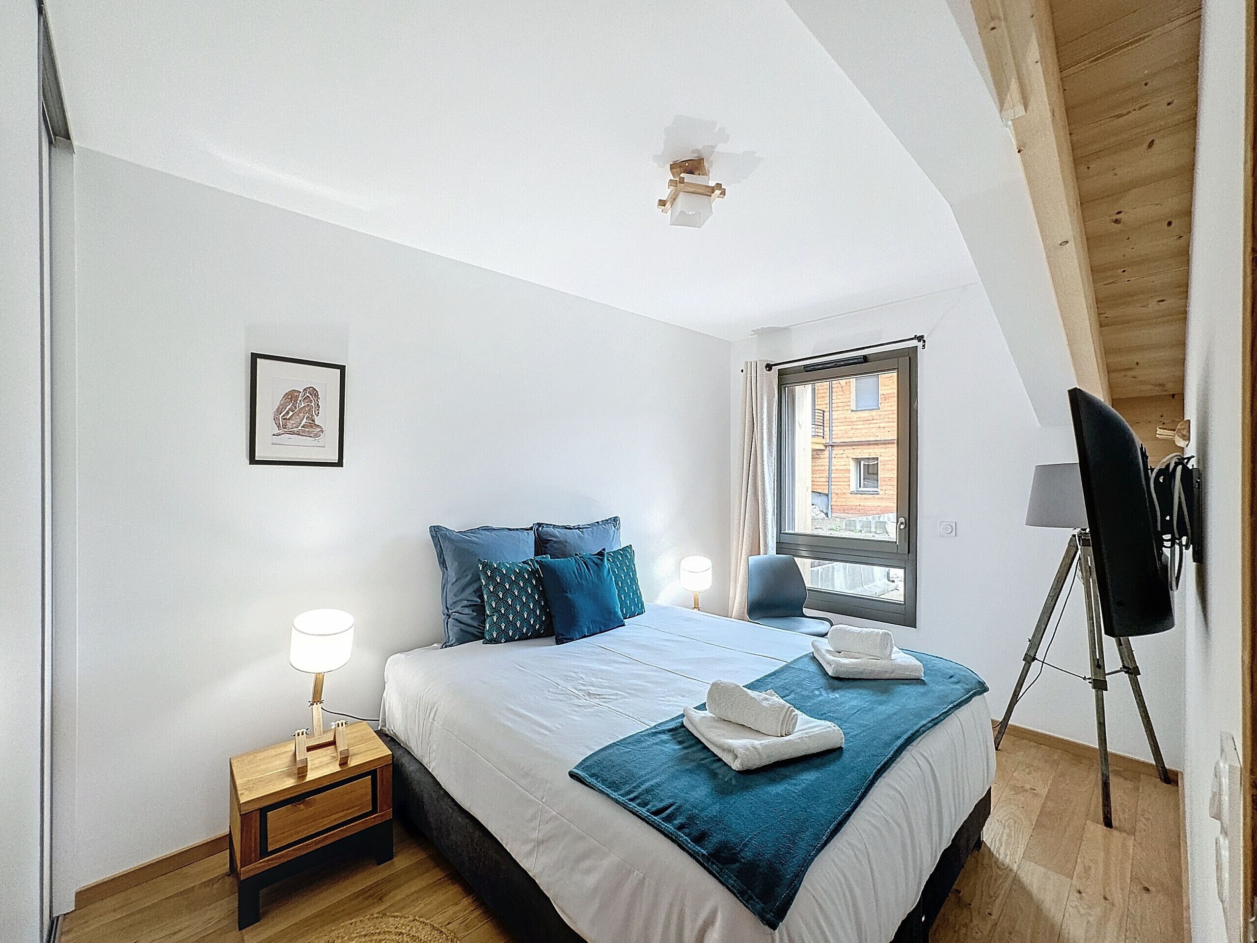 A bedroom with a bed, a bedside table with a lamp, a window with a view of buildings, and a ceiling light fixture.