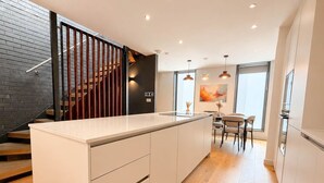 A modern kitchen with a central island, wooden flooring, and a staircase with a black railing.