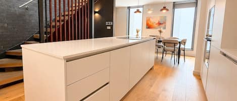 A modern kitchen with a central island, wooden flooring, and a staircase with a black railing.