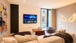 A modern living room with a sofa, a coffee table, a bean bag, a flat-screen TV displaying Disney channels, and a wall with a neon wing decoration.