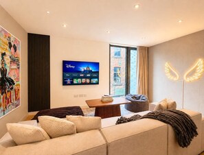 Moderni olohuone, jossa on sohva, sohvapöytä, säkkituoli, litteänäyttöinen televisio, jolta näkyy Disneyn kanavia, ja seinä, joka on koristeltu neonvaloin muodostetulla siipikoristeella.