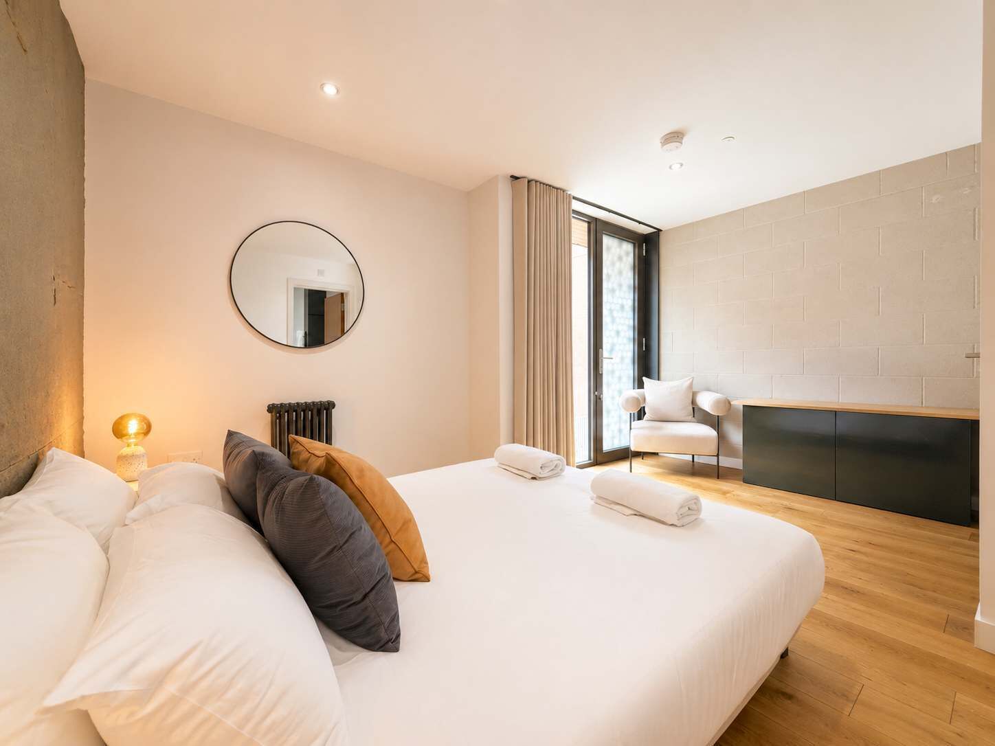 A modern bedroom with a large bed, a round mirror, a fireplace, and a view of the outdoors.