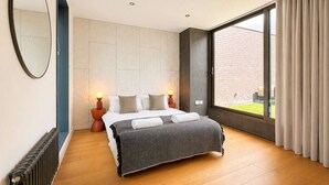 A modern bedroom with a large bed, a round mirror, a radiator, and a view of a brick building through a window.