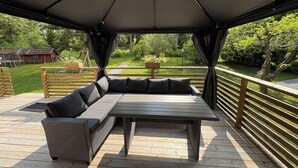 Un espacio de estar al aire libre cubierto, con una mesa y bancos, rodeado por una terraza de madera y vistas a un jardín y un edificio rojo al fondo.
