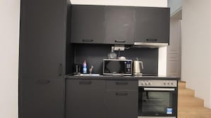 Eine kleine Küche mit Kühlschrank, Mikrowelle, Backofen und einer Arbeitsfläche mit Wasserkocher und Flasche.