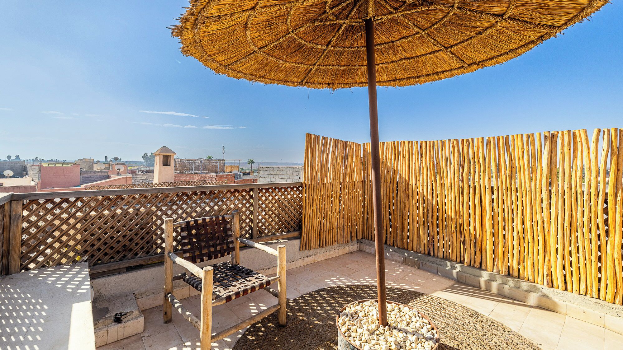 Eine Dachterrasse mit einem Holzstuhl, einem gewebten Sonnenschirm und einem Zierzaun.