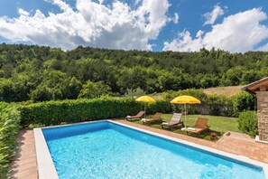 Una piscina rectangular con agua azul cristalina, rodeada por un patio de ladrillo. Hay dos sillas reclinables con sombrillas amarillas, y la piscina está situada frente a un fondo de árboles verdes exuberantes y un cielo azul despejado.