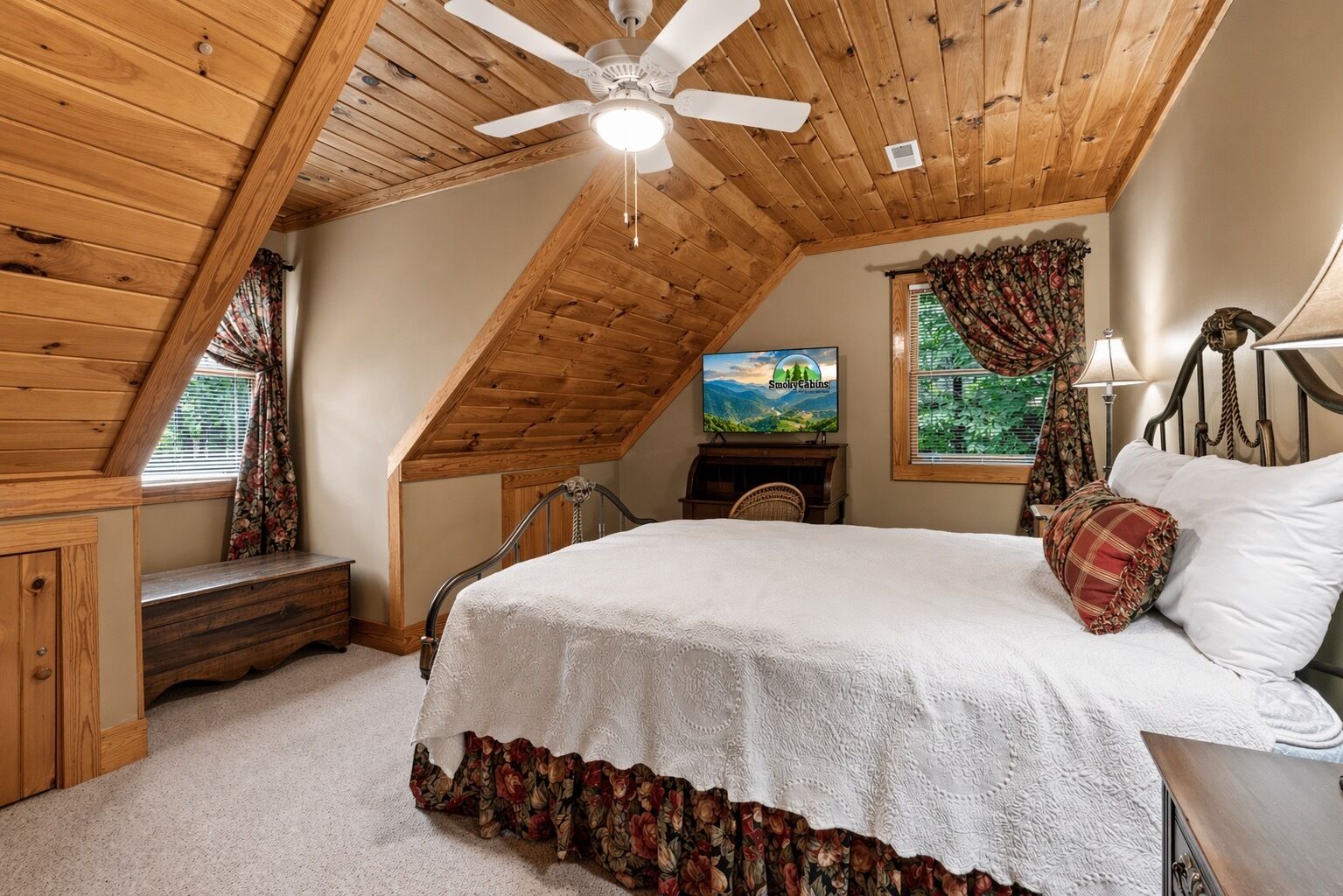 Un dormitorio con techo inclinado, una cama con colcha floral, un televisor y una ventana con cortinas.