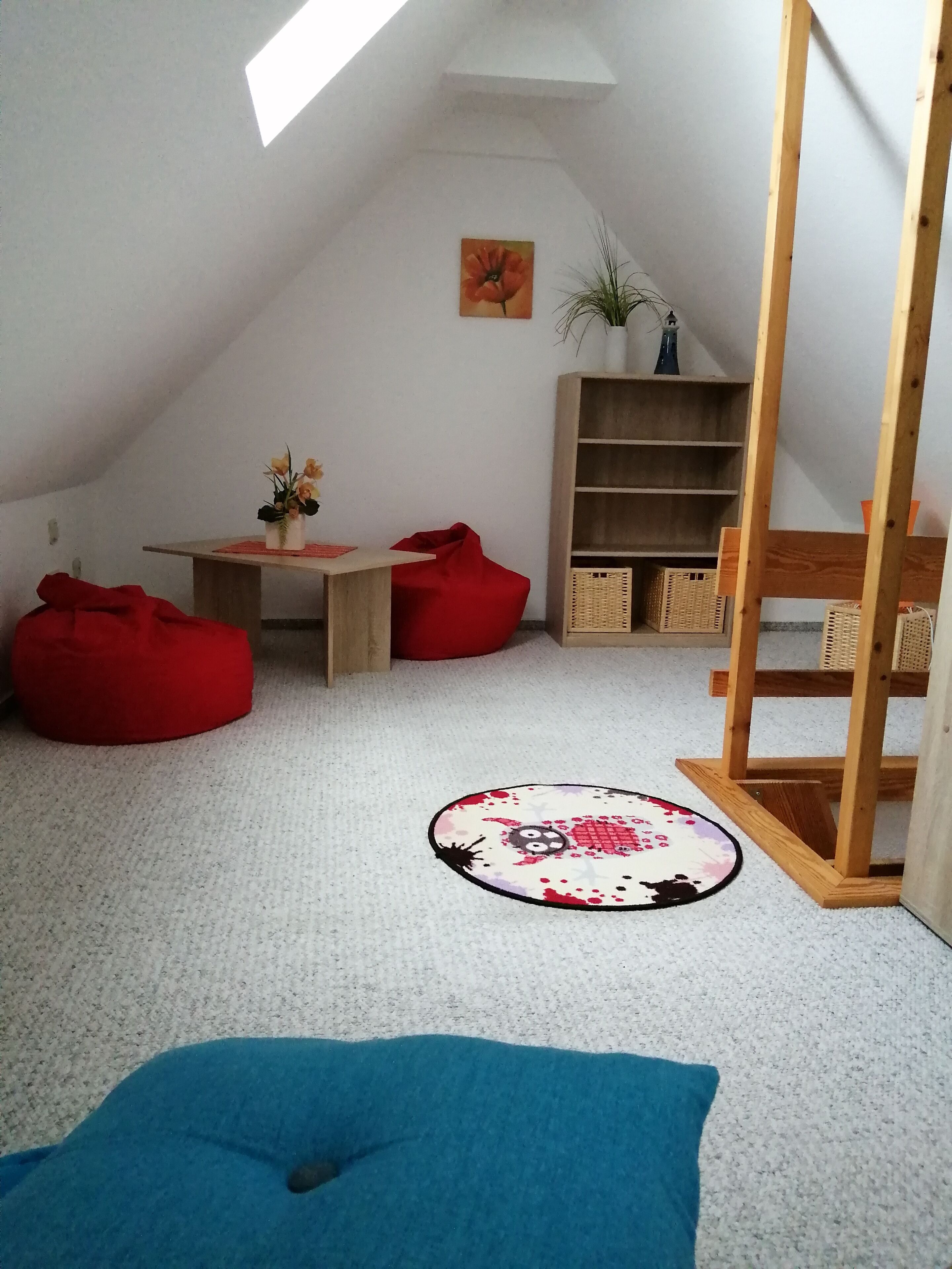 A cozy, well-lit attic room with a sloped ceiling, a wooden desk, a red bean bag, a blue cushion, a small round rug, and a wooden bookshelf.
