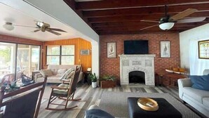 A cozy living room with a fireplace, wooden beams, and a large window. - Lakefront Bliss | Kayaks • Paddleboards • Fire Pit • Snowbird Specials (Melrose)