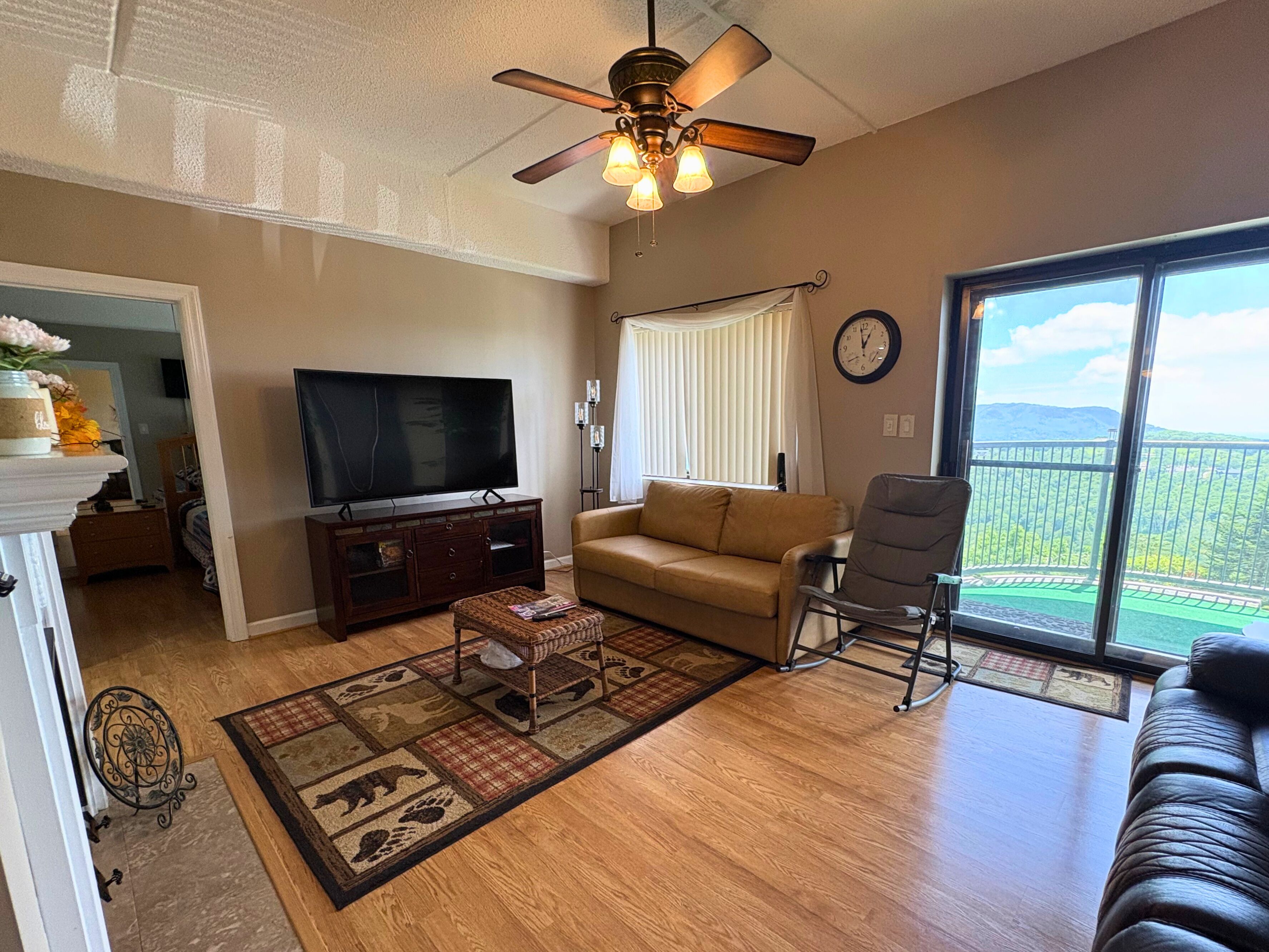 A living room with a television, sofa, armchair, and a view of a green landscape through large windows.