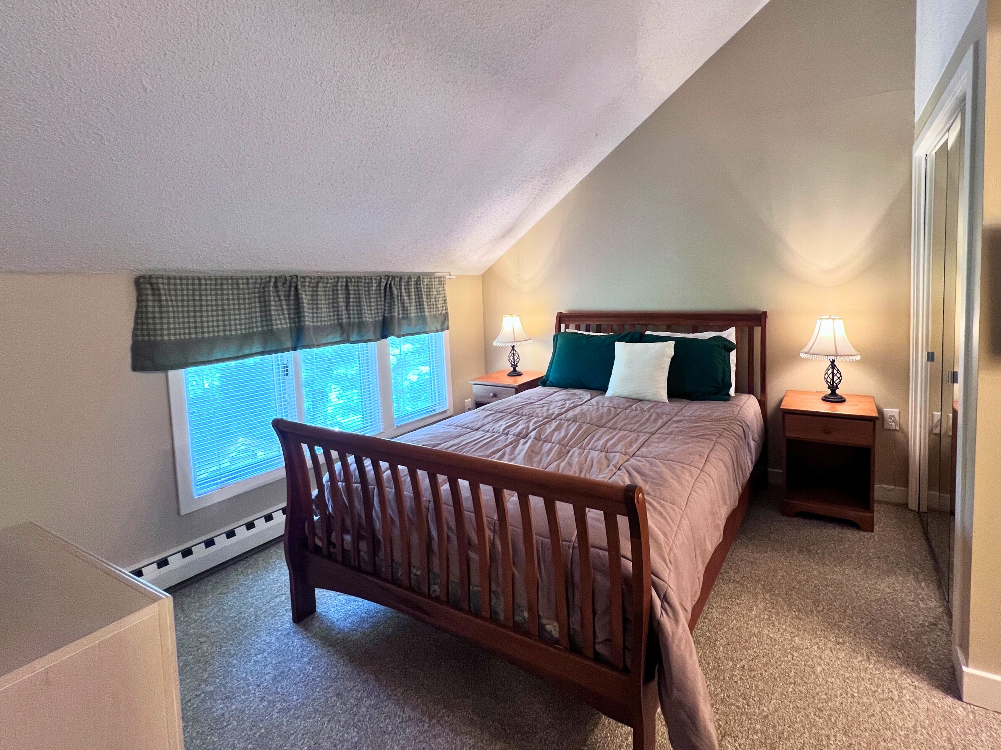 A bedroom with a sloped ceiling, a window with blinds, a bed with a wooden headboard, two bedside tables with lamps, and a carpeted floor.
