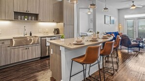 A modern kitchen with wooden cabinets, a central island with bar stools, pendant lighting, and a dining area with a blue sofa and coffee table. - Luxurious New Unit|2Bd2Ba|Pool, Golf Sim (Austin)