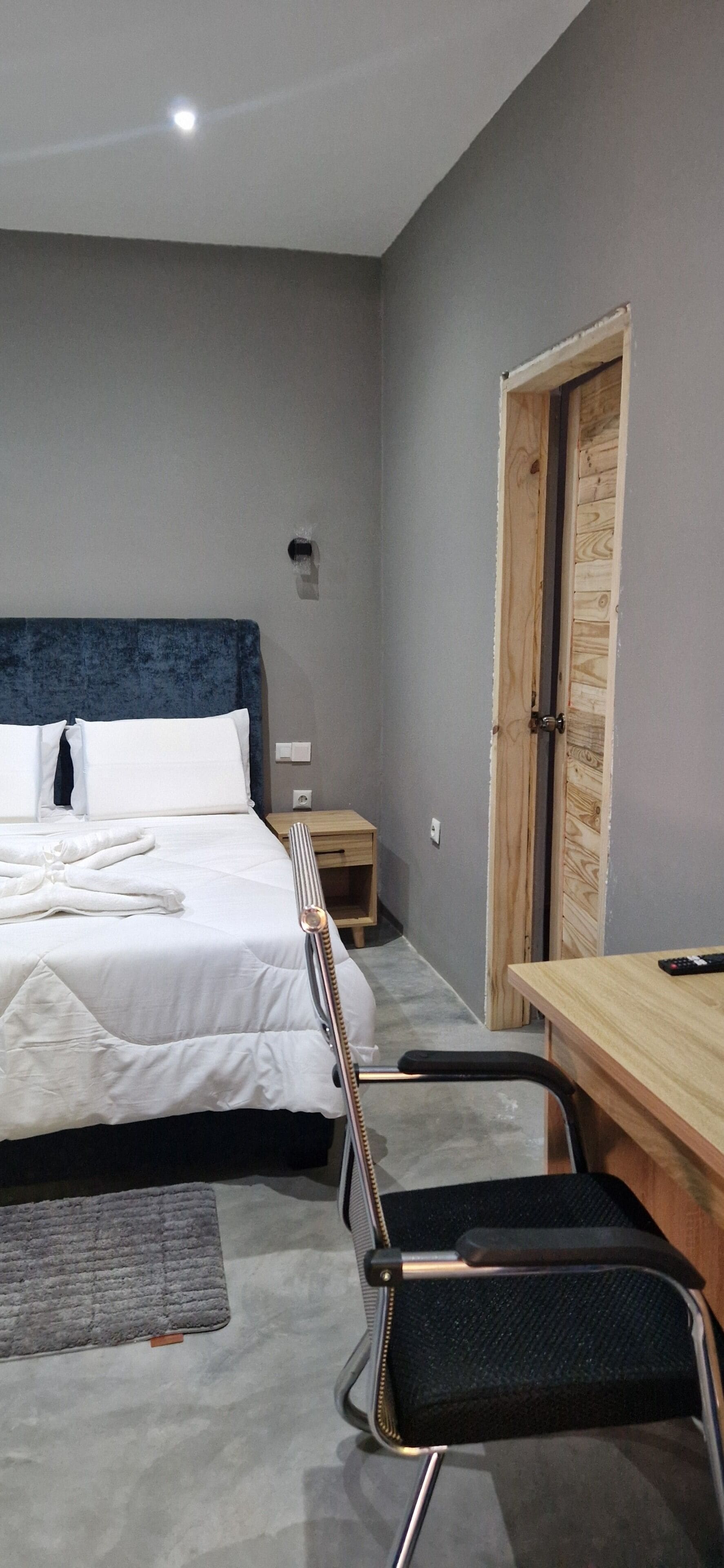 A bedroom with a bed, a desk, a chair, a wooden door, and a small nightstand.