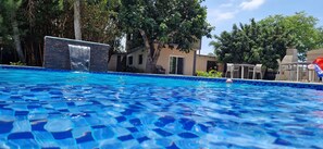 A swimming pool with blue mosaic tiles, surrounded by trees and a building with a waterfall feature.
