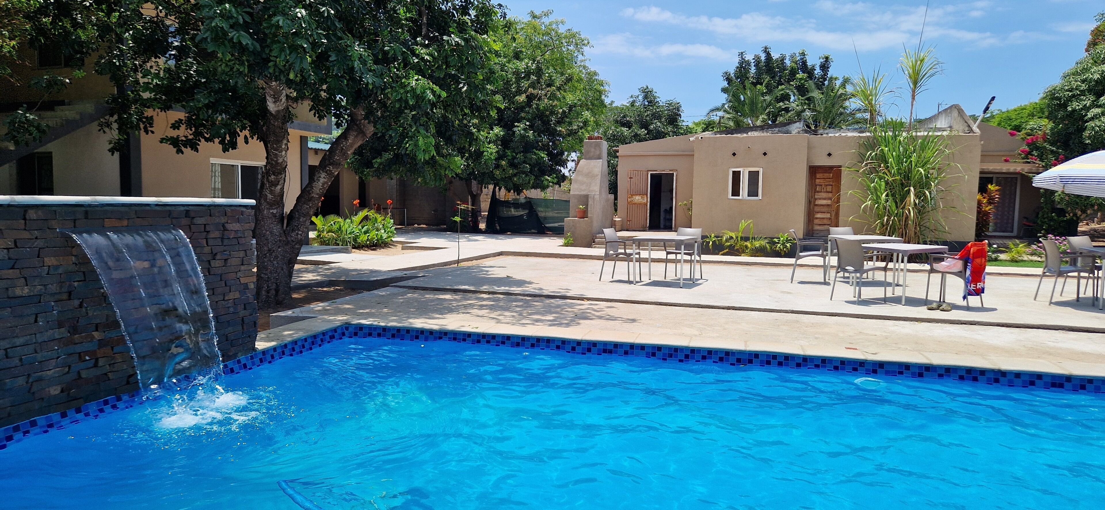 Una piscina con una cascada, rodeada de mobiliario exterior y un edificio con puerta y ventanas.