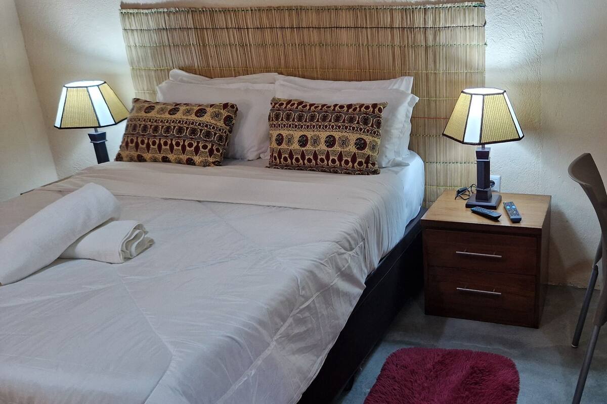 A neatly made bed with white linens and patterned pillows, a bedside table with a lamp, a wooden desk, and a chair.