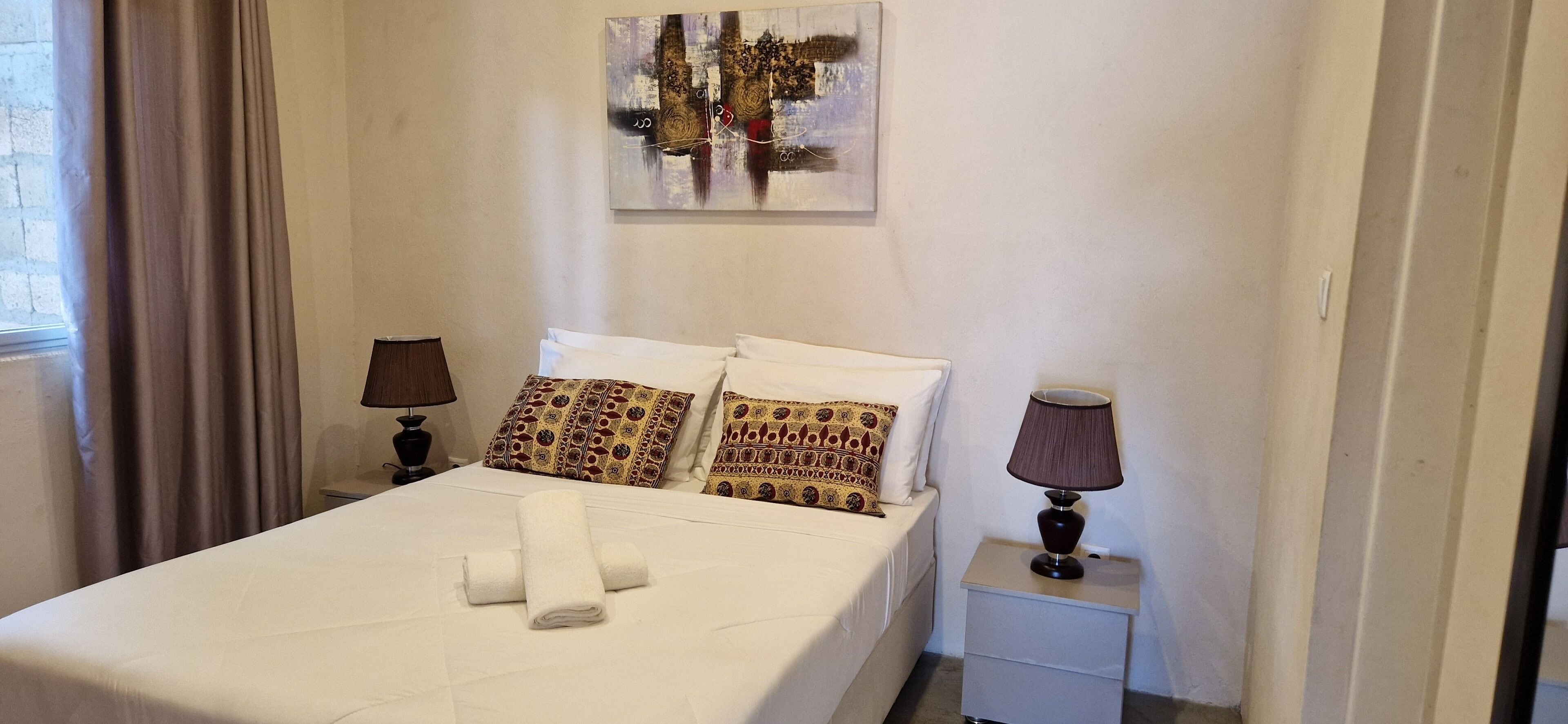 Una cama bien hecha con sábanas blancas y almohadas estampadas, una lámpara de noche y un cuadro en la pared.