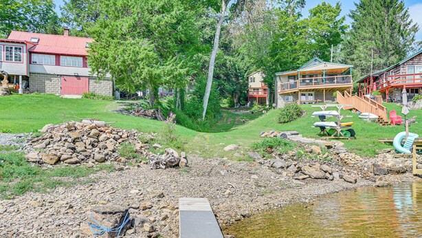 A lakeside property with a dock leading to a small boat shed, surrounded by lush greenery and a stone wall.