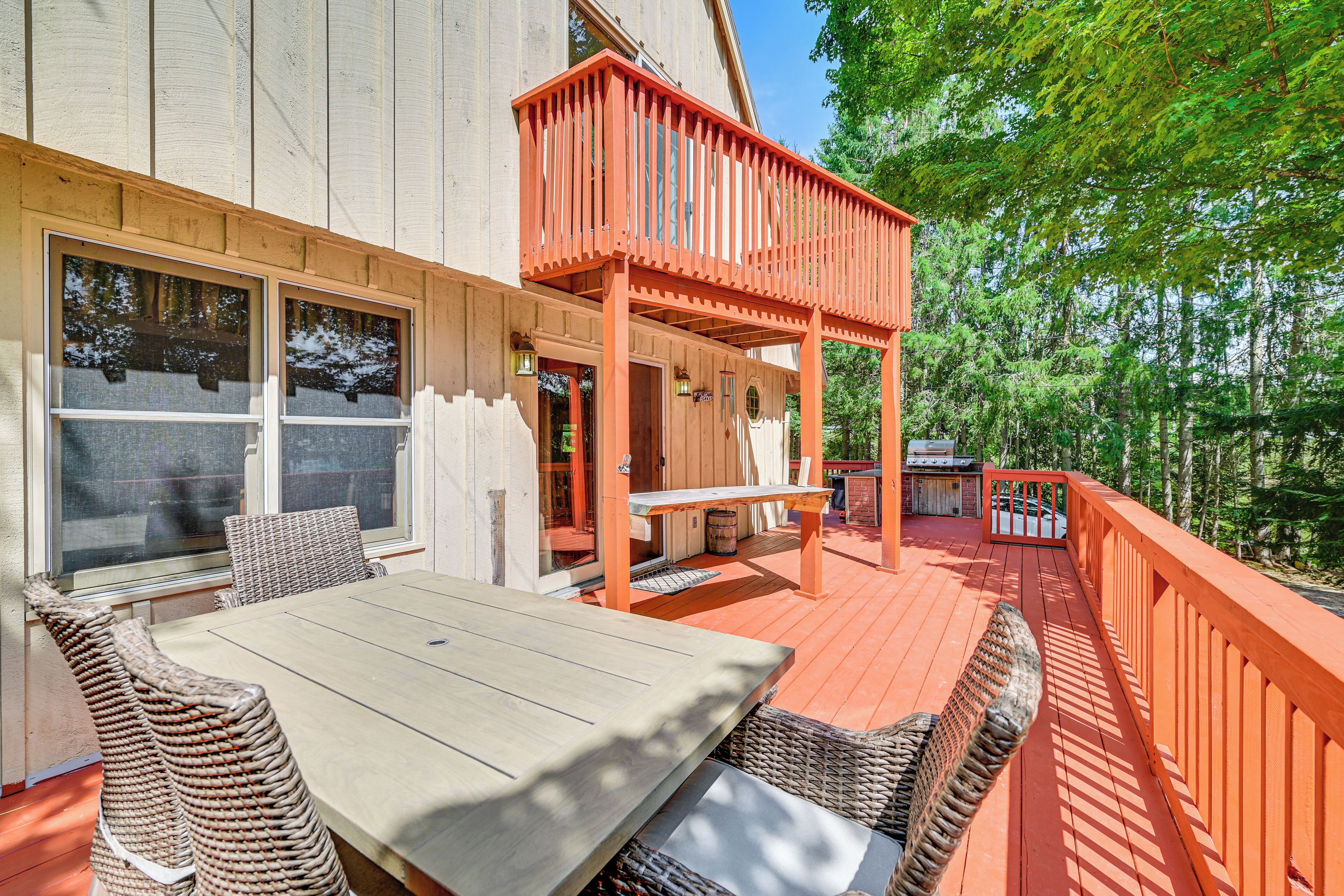 A wooden deck with a dining table and chairs, a covered outdoor seating area, and a view of trees.