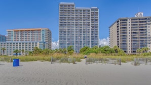 Ein Strandhotel mit mehreren Hochhäusern, einem Sandstrand und einer Hecke, die Strand und Hotellandschaft voneinander abgrenzt.