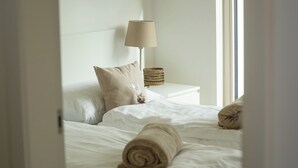 Un lit bien fait, avec une serviette enroulée, une lampe de chevet et un panier tressé posé sur une table de chevet blanche.