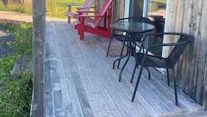 A wooden deck with red Adirondack chairs and a round table, overlooking a grassy field.