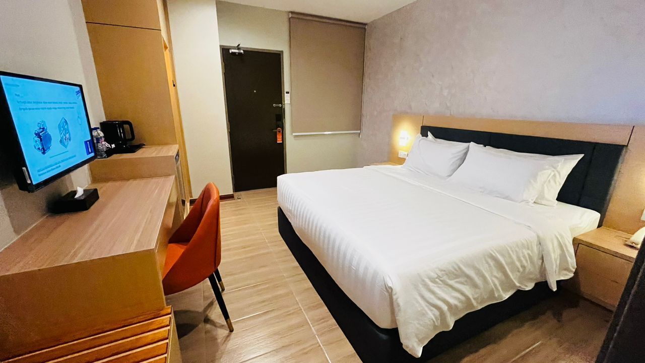 A hotel room with a bed, a desk with a computer, a chair, and a bedside table with a lamp.