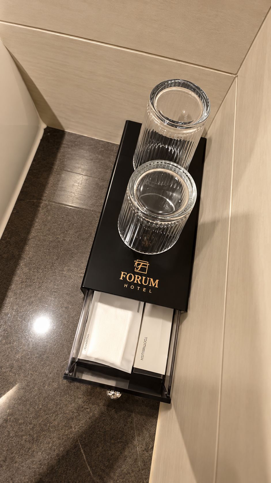Un minibar negro con dos vasos de vidrio transparentes y una botella de agua, situado en un rincón de una habitación con piso de baldosas.