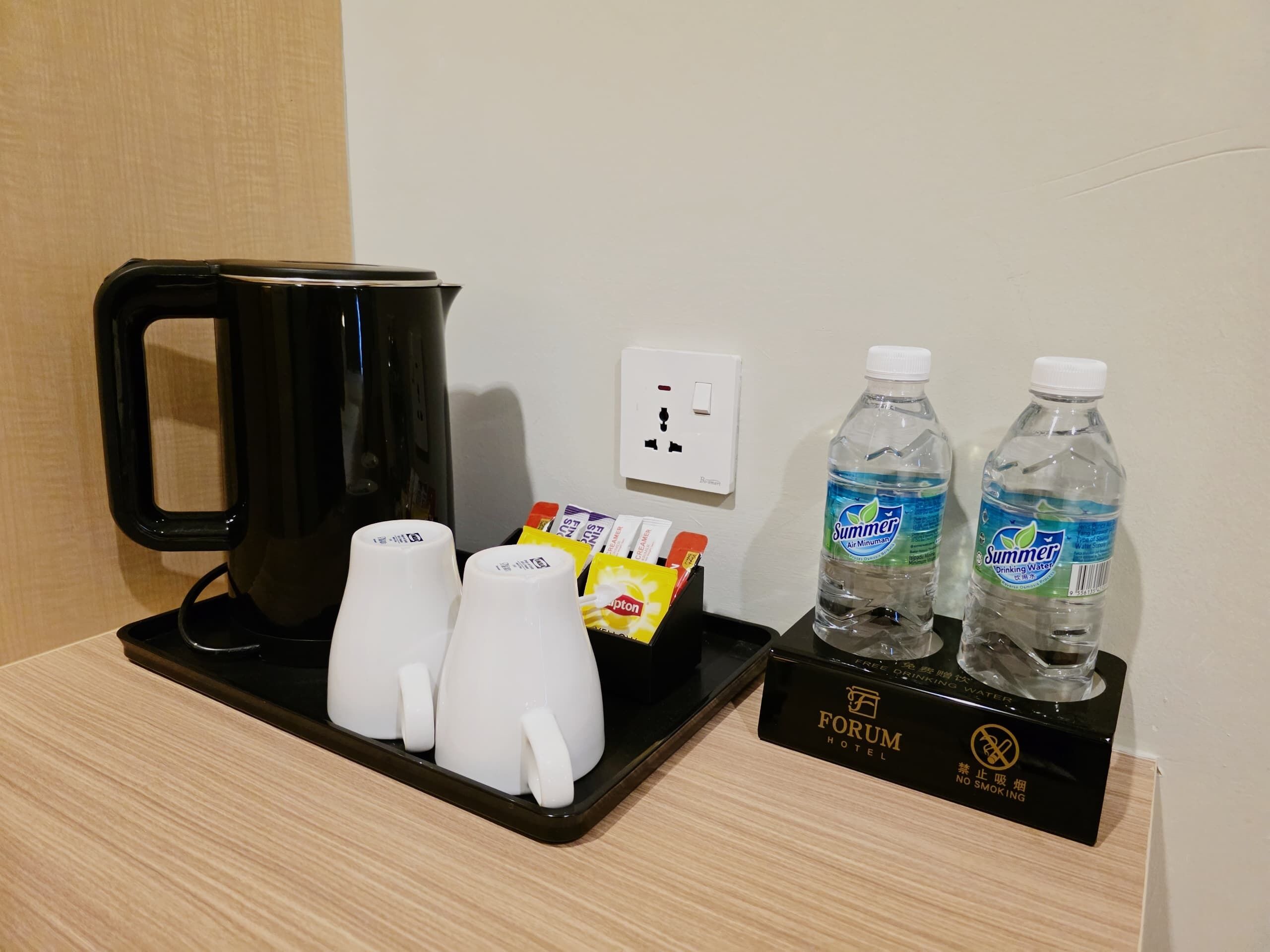 Minibar på et hotelværelse med en sort kaffemaskine, to hvide kopper, en bakke med tissues, to vandflasker og et 'Rygning Forbudt' skilt.