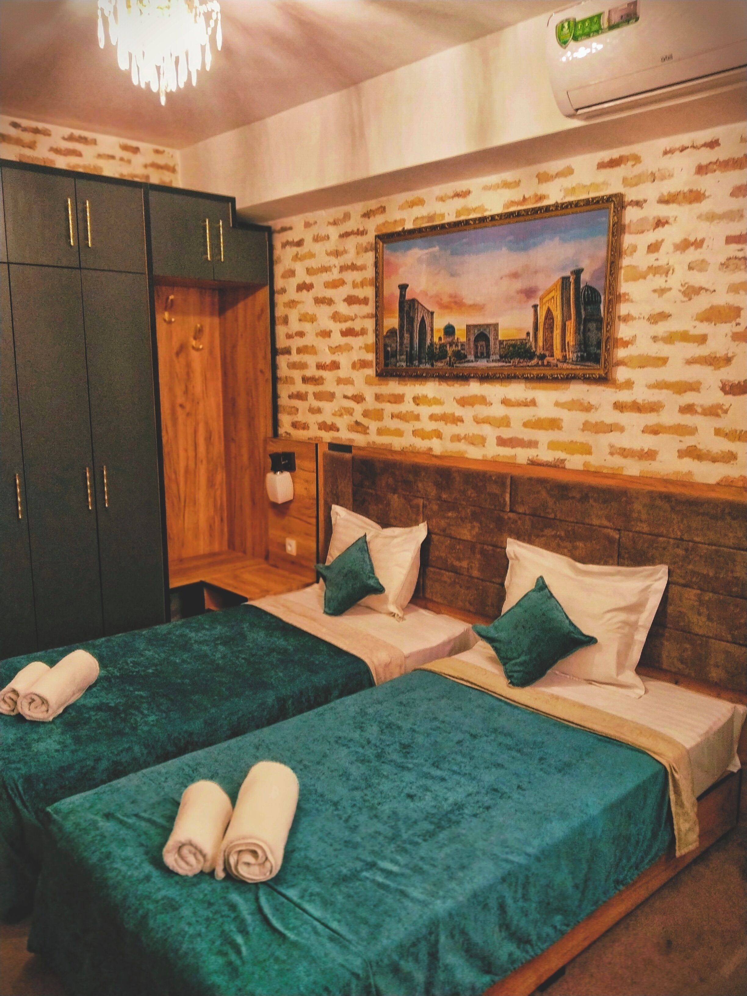 A hotel room with two beds, a wardrobe, a painting on the wall, and an air conditioning unit.