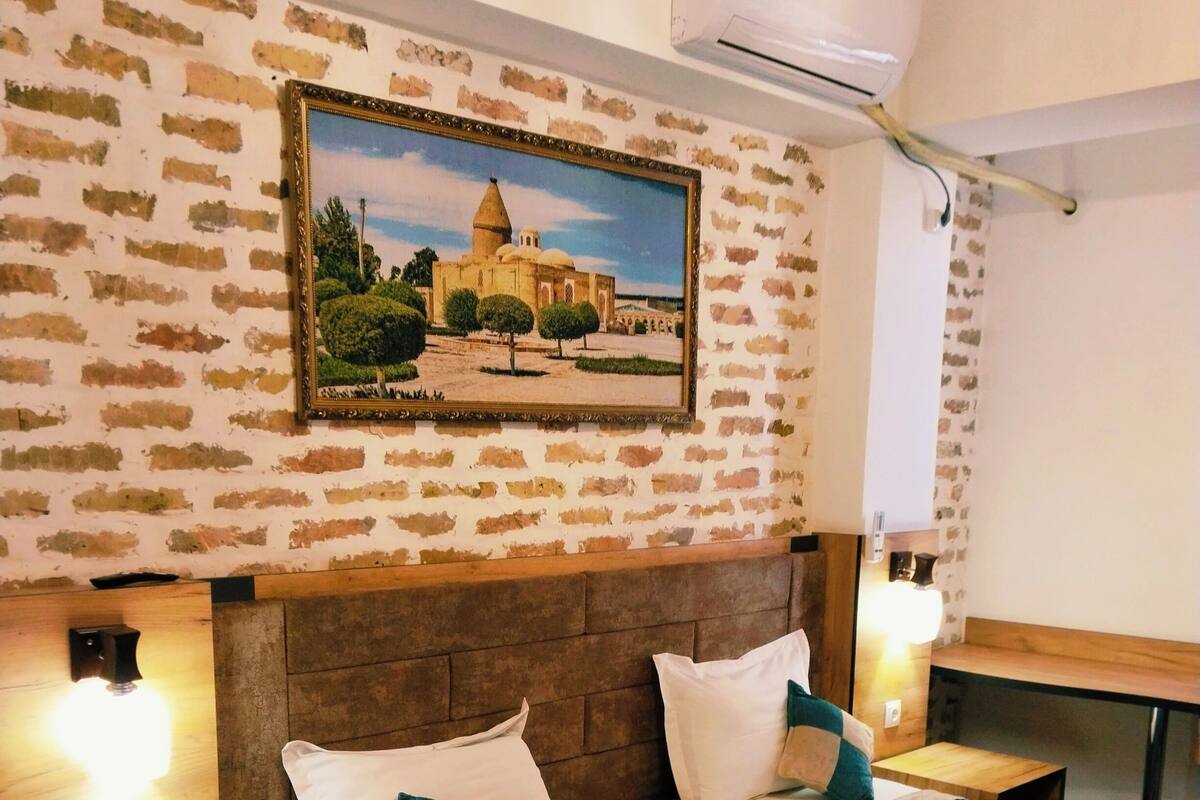 Une chambre d’hôtel avec un mur décoré d’un motif en briques, un grand lit avec une literie de couleur turquoise, deux tables de chevet avec des lampes et un tableau représentant un bâtiment.