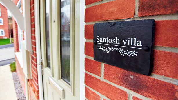 Ein schwarzes Schilt aus Schiefer mit der Aufschrift "Santosh Villa", angebracht an einer roten Ziegelwand.