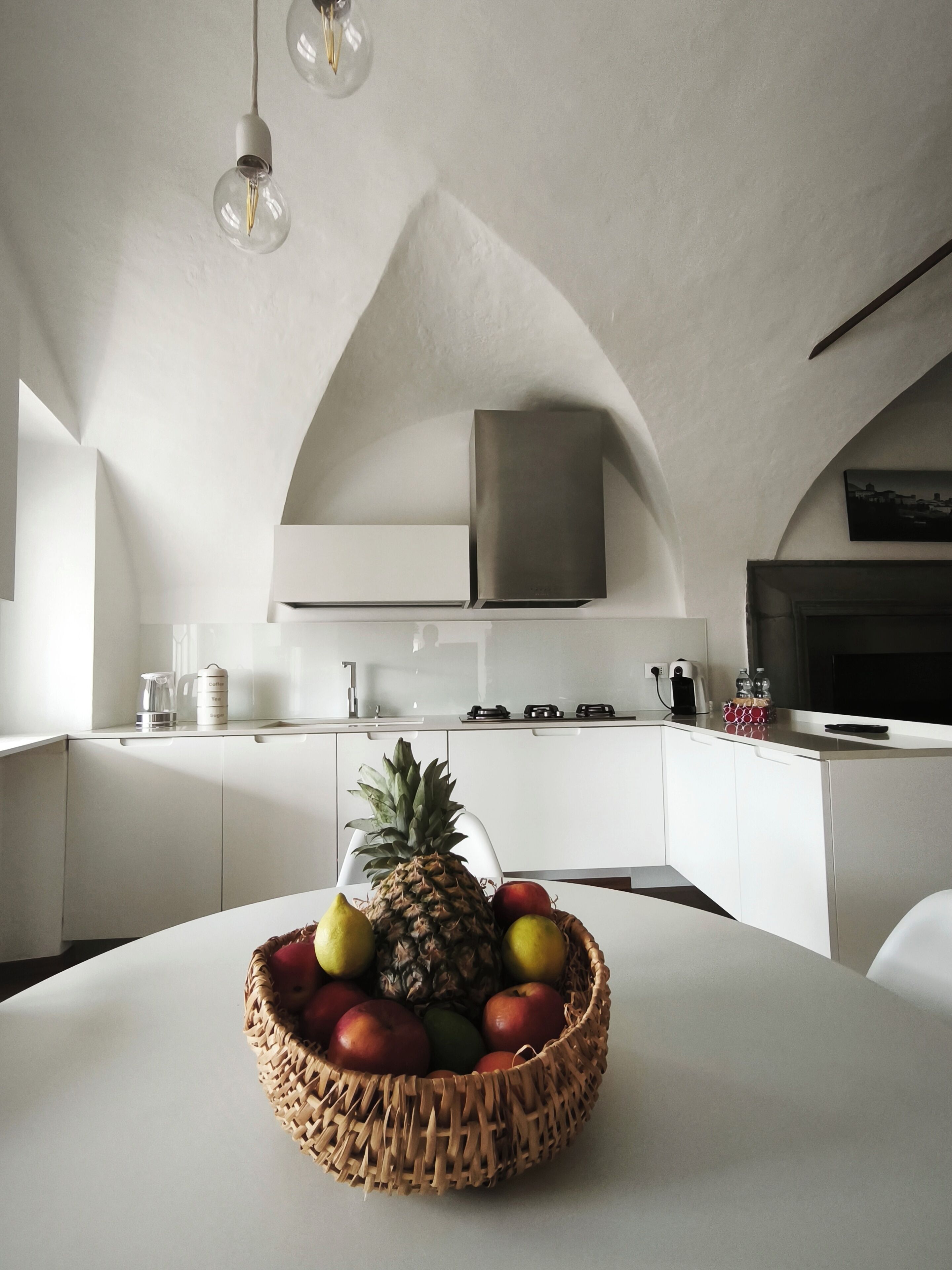 Une cuisine moderne avec des meubles blancs, une hotte de cuisinière en acier inoxydable et un panier de fruits sur la table.