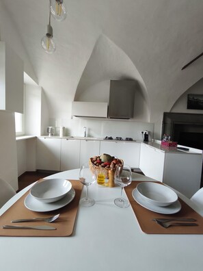 A modern kitchen with white cabinets, a dining table set with plates, glasses, and cutlery, and a fruit basket on the counter.
