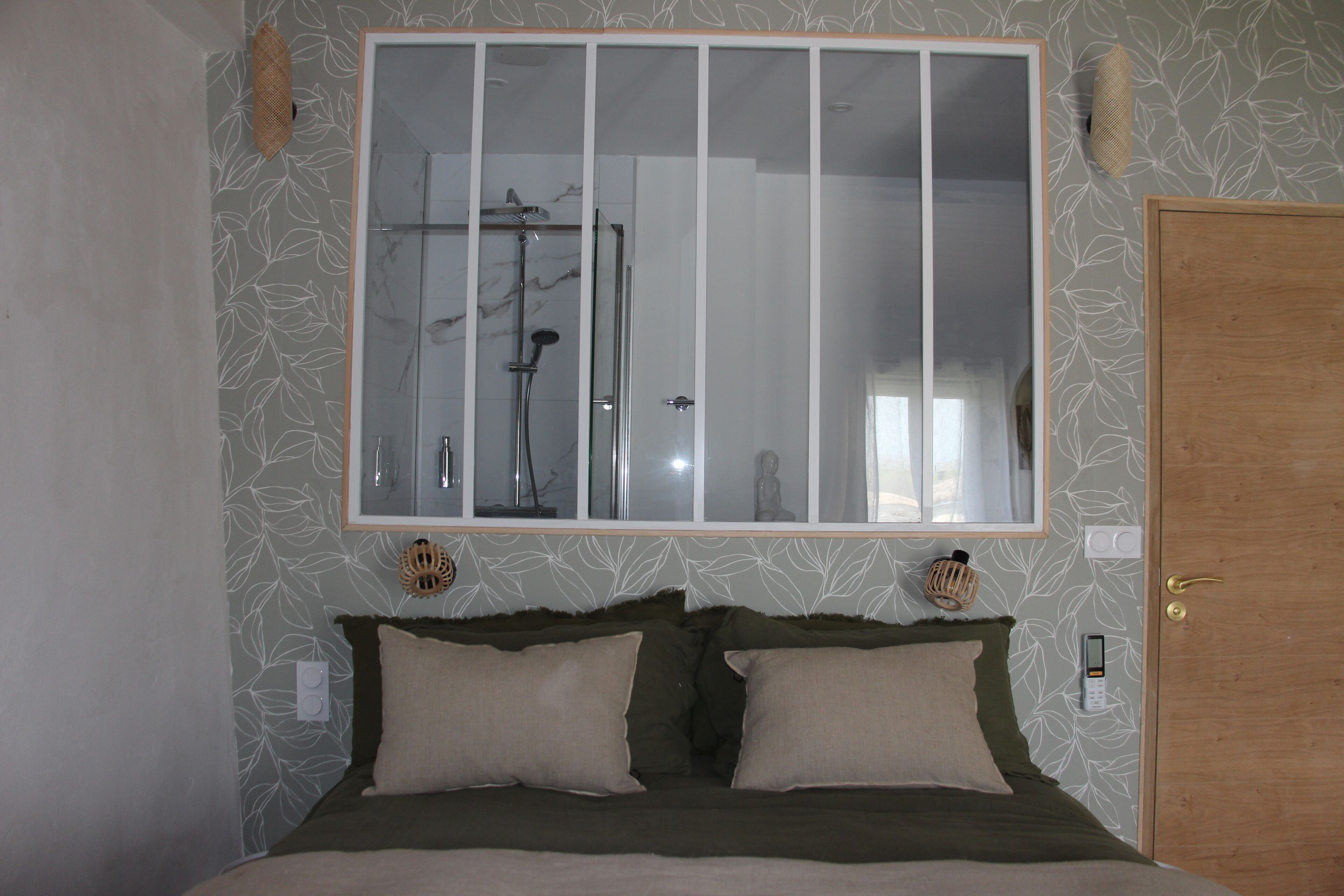 A bedroom with a large mirror, a bed with two pillows, a wooden door, and a shower visible through the mirror.