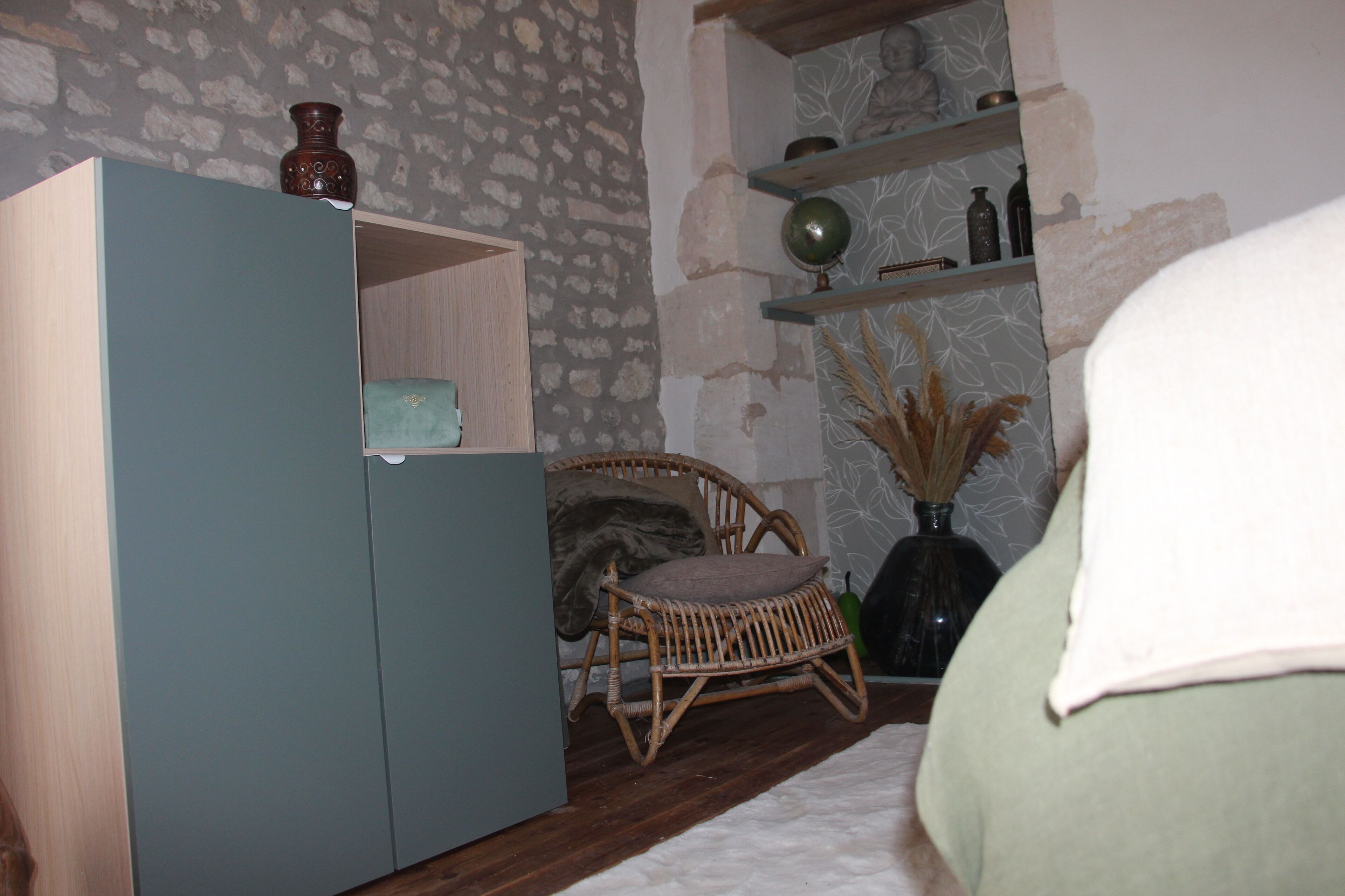 A room with a wooden wardrobe, wicker chair, and shelves with decorative items.