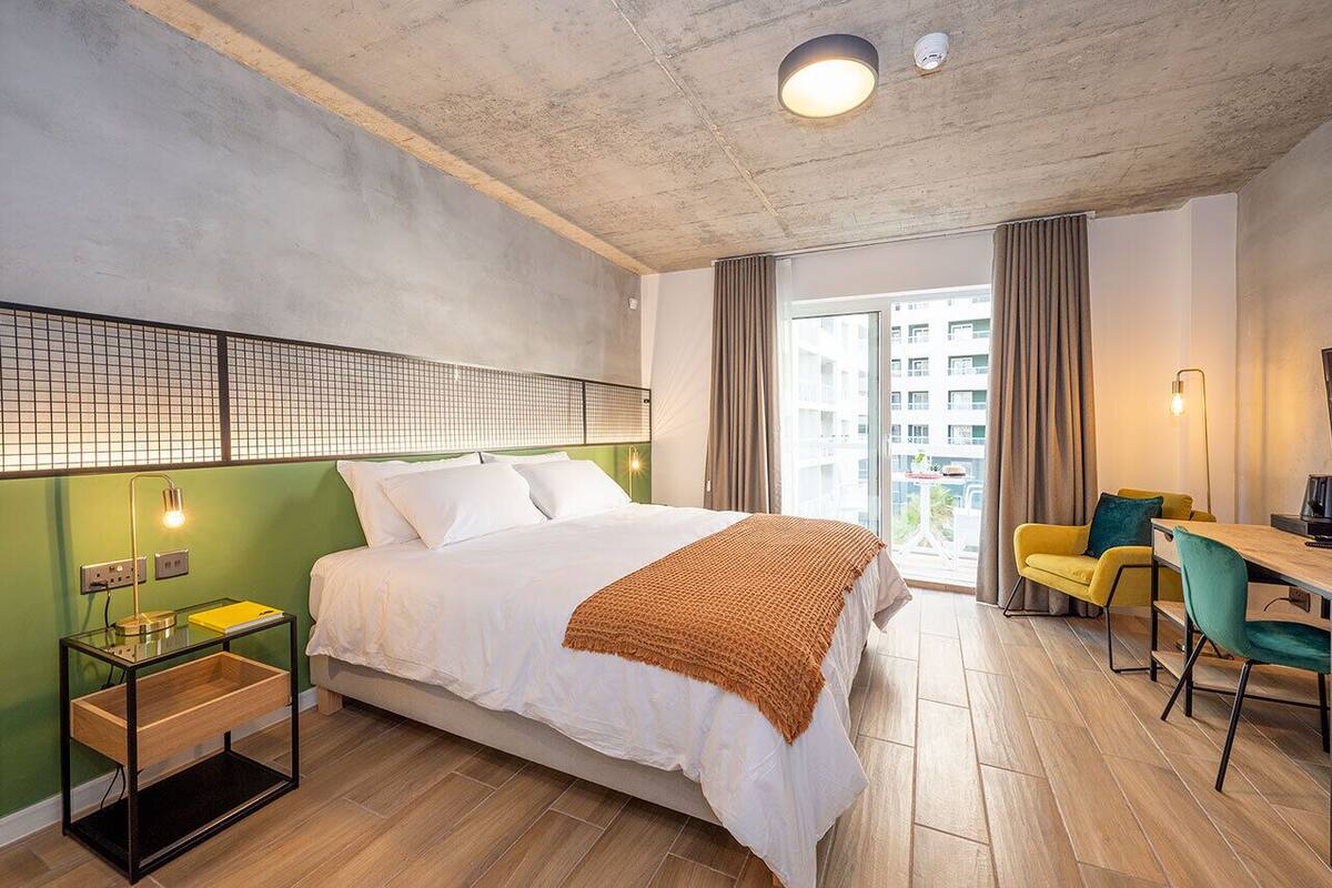 Ein modernes Hotelzimmer mit einem großen Bett, einem Schreibtisch mit Stühlen, einem kleinen Tisch mit Lampe und Blick auf die Stadt.
