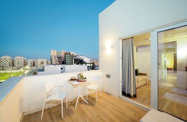 A balcony with a table and chairs, a sliding glass door leading to an interior room, and a view of a cityscape at dusk. - Fort40 Guesthouse (Sliema)