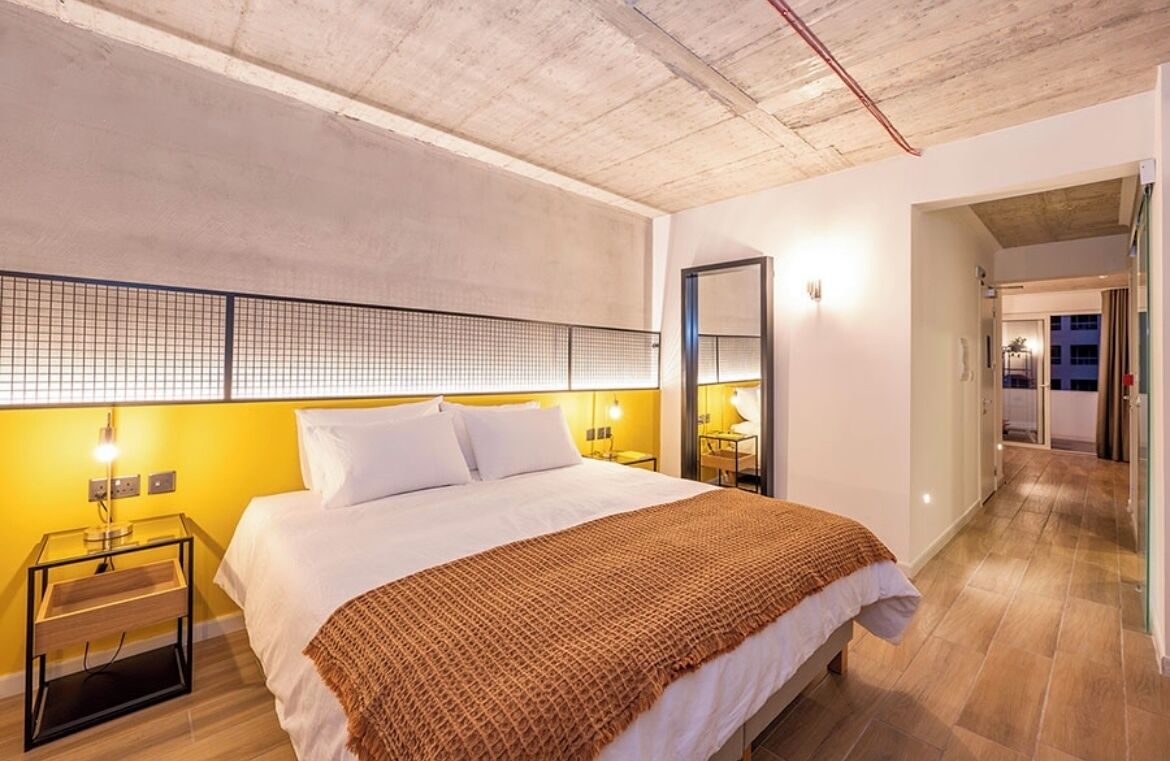 A modern hotel room with a large bed, a wooden floor, a yellow accent wall, and a visible hallway leading to other rooms.