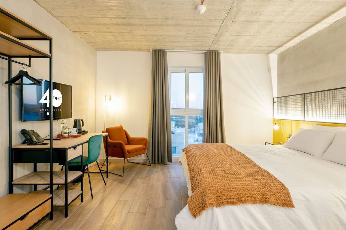Ein Hotelzimmer mit einem Bett, einem Schreibtisch mit Computer, einem Stuhl und einem Fenster mit Vorhängen.