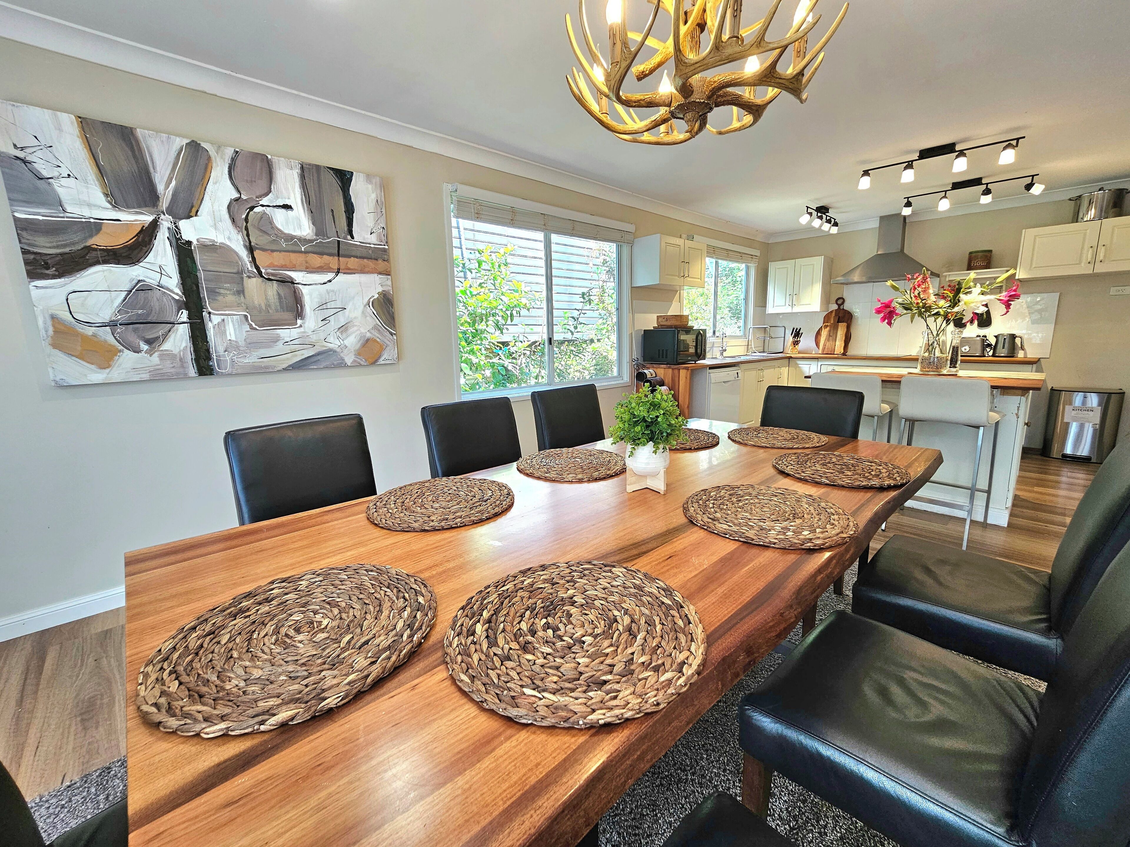 A modern dining room with a wooden table, woven placemats, black leather chairs, and a chandelier.