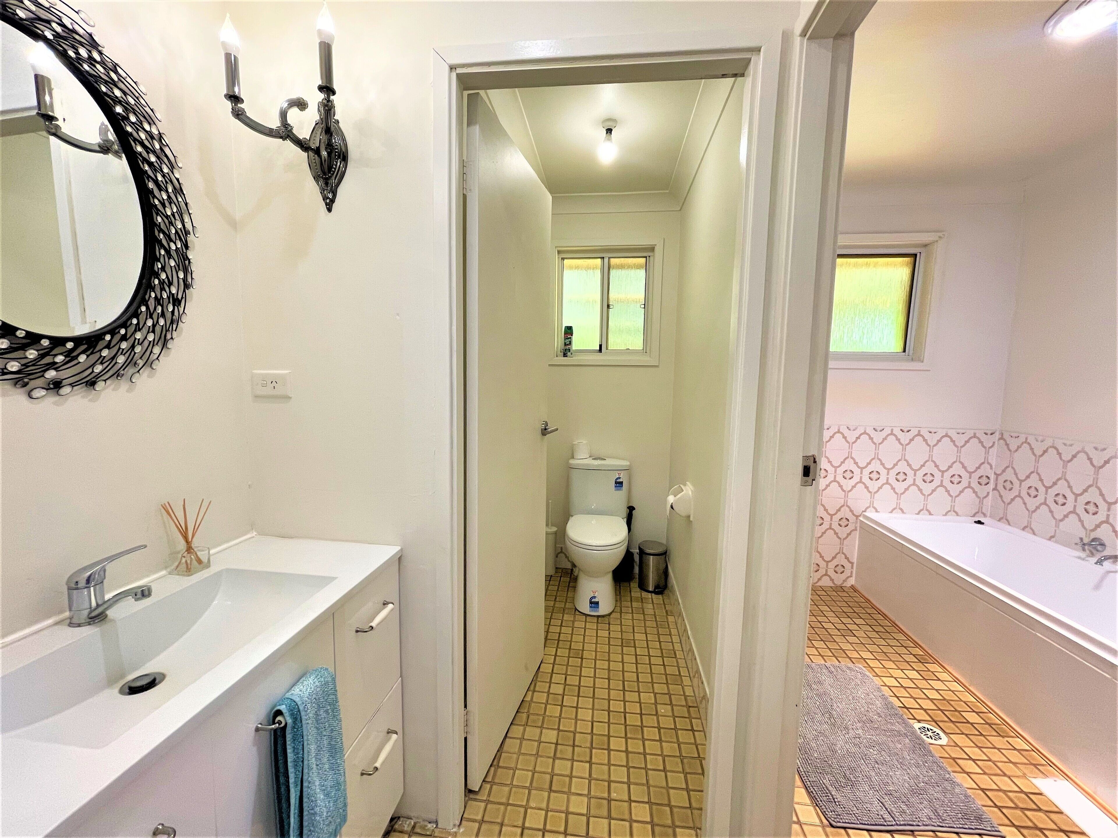 Un baño con lavamanos blanco, espejo y inodoro. Hay una ventana con cortinas, una bañera y un papel tapiz decorado.