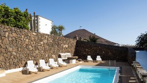 Ein Poolbereich mit weißen Liegestühlen, einer Steinmauer und einem Berg im Hintergrund.