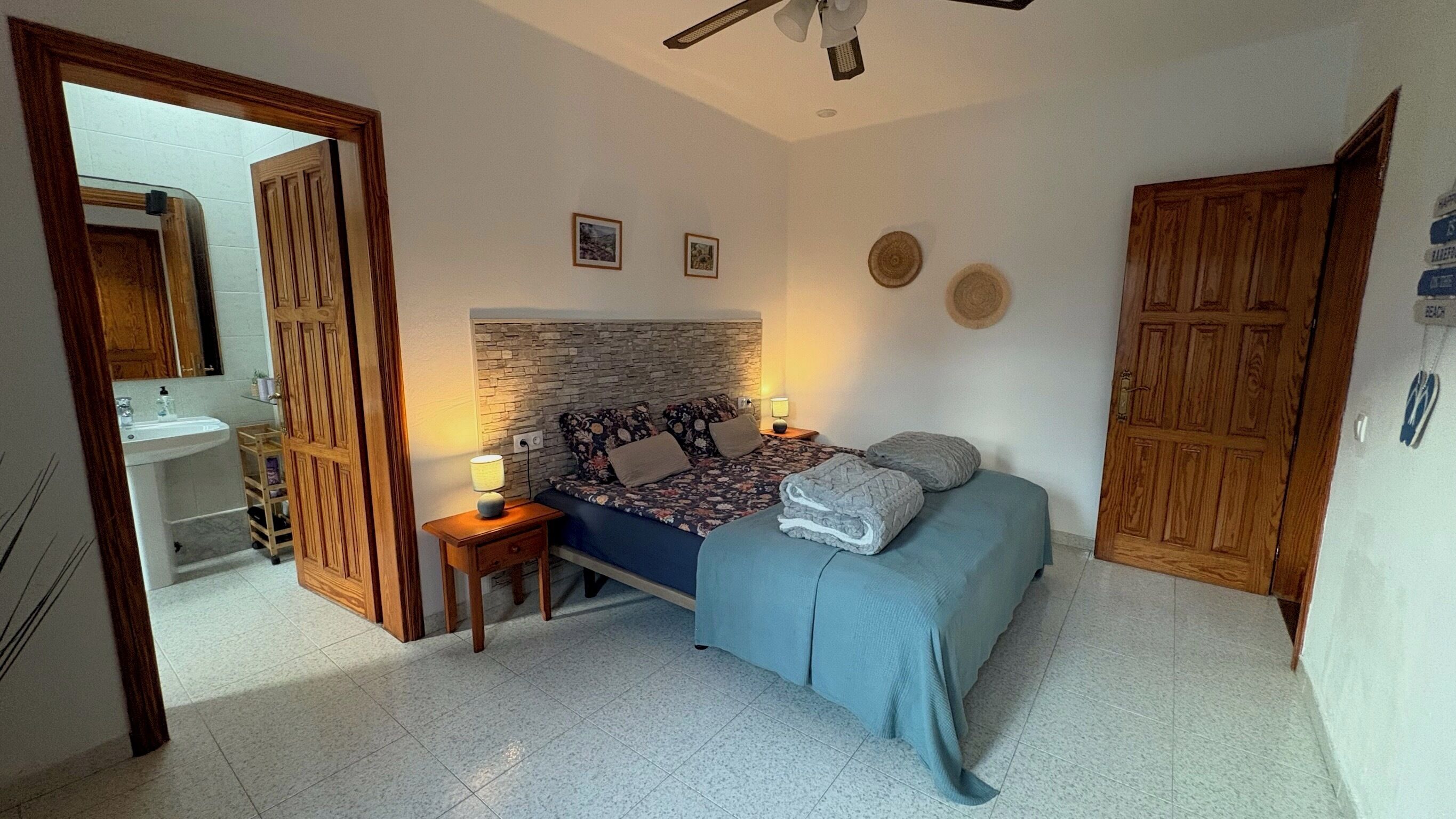A bedroom with a bed, bedside tables, a nightstand, a mirror, a wooden wardrobe, a bathroom with a sink, and a tiled floor.