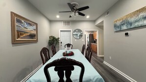 A dining room with a long table, chairs, a ceiling fan, a painting, and a kitchen in the background.